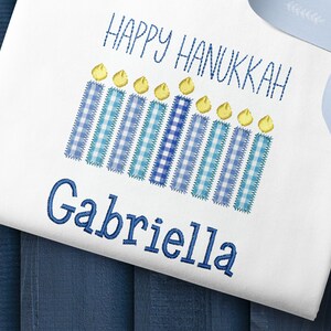 May include: White onesie with blue and white checkered candles and the text "Happy Hanukkah" and the name "Gabriella" embroidered on it.
