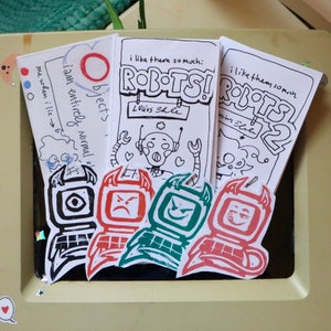 以下が含まれることがあります： ロボットをテーマにしたイラストとテキストの手作りアート作品のコレクション。アートワークには、ロボットの絵、「ROBOTS!」と「ROBOTS 2」のテキスト、手書きのメモが含まれています。カラーパレットは黒、赤、緑です。