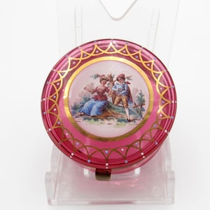 May include: A round, deep pink glass trinket box featuring a central circular medallion with a painted scene of two figures in a landscape. The lid is decorated with gold scalloped lines and small white and blue dots around the perimeter.