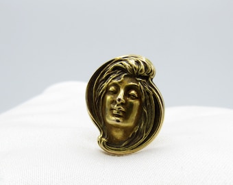 May include: A gold-toned metal ring featuring a detailed design of a woman's face. The woman's hair is flowing and her eyes are closed.