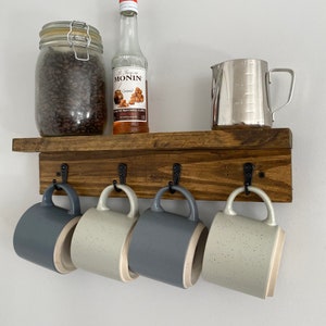 May include: A rustic wooden shelf with five hooks for hanging mugs. Four mugs are hanging on the hooks, two are grey and two are white with black speckles. A glass jar filled with coffee beans and a bottle of caramel syrup are on the shelf.