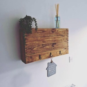 May include: A rectangular wooden wall shelf with a natural grain finish, featuring three black metal hooks along the bottom edge. A small grey house-shaped sign hangs from the centre hook with the text A home without a cat is just a house.
