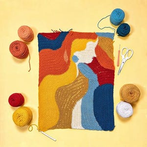 May include: A rectangular crochet project featuring abstract wavy shapes in orange, yellow, cream, red, and blue yarn. The piece is surrounded by several balls of matching yarn, a pair of silver scissors, and two crochet hooks on a yellow surface.
