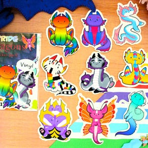 Könnte beinhalten: Eine Sammlung farbenfroher Drachenaufkleber mit verschiedenen Designs, darunter Regenbogen-, gestreifte und geflügelte Drachen. Ein Aufkleberpaket mit der Aufschrift "Pride Dragons" ist ebenfalls sichtbar, zusammen mit dem Wort "Vinyl". Die Aufkleber sind auf einer Holzoberfläche angeordnet.