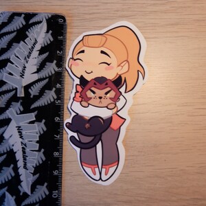 Puede incluir: Una pegatina con un personaje de dibujos animados con cabello rubio y un atuendo rosa y naranja que sostiene un gato negro y rojo. El personaje tiene una expresi&oacute;n feliz y sonr&iacute;e.