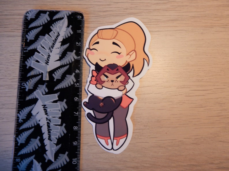 Puede incluir: Una pegatina con un personaje de dibujos animados con cabello rubio y un atuendo rosa y naranja que sostiene un gato negro y rojo. El personaje tiene una expresi&oacute;n feliz y sonr&iacute;e.