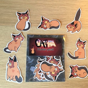 Puede incluir: Un paquete de pegatinas con diferentes ilustraciones de gatos. Las pegatinas son todas de estilo de dibujos animados y presentan gatos con máscaras rojas y negras. Las pegatinas están dispuestas en un círculo alrededor de una tarjeta que dice "Cat-Ra Sticker Pack".