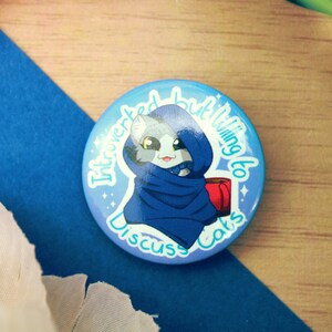 May include: A blue and white button with a cartoon cat wearing a blue hooded robe and holding a red mug. The text on the button reads "Introverted but willing to discuss cats".