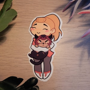 Puede incluir: Una pegatina con un personaje de dibujos animados con cabello rubio y un gato rojo y negro. El personaje sonr&iacute;e y sostiene al gato en sus brazos. La pegatina est&aacute; sobre una superficie de madera.