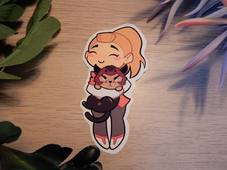 Puede incluir: Una pegatina con un personaje de dibujos animados con cabello rubio y un gato rojo y negro. El personaje sonr&iacute;e y sostiene al gato en sus brazos. La pegatina est&aacute; sobre una superficie de madera.