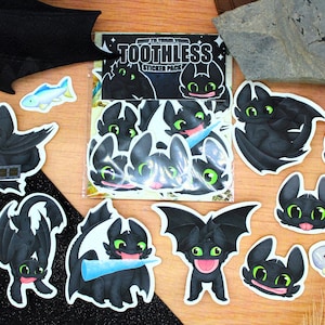 Puede incluir: Un conjunto de pegatinas de Toothless de la película "Cómo entrenar a tu dragón". Las pegatinas muestran al dragón negro con ojos verdes en varias poses, junto con peces azules. También se incluye un paquete de pegatinas con el texto "Toothless".
