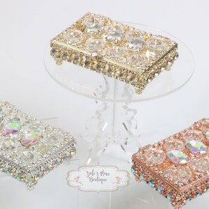 Puede incluir: Tres cajas decorativas con tapas transparentes y bordes adornados con joyas. Las cajas son doradas, plateadas y de oro rosa. Cada caja tiene una tapa transparente y está adornada con diamantes de imitación iridiscentes.