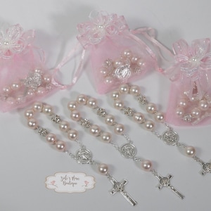 Puede incluir: Tres rosarios de perlas rosas con cruces y medallas de plata, empaquetados en bolsas de organza rosa con ribete de encaje blanco y una flor blanca.