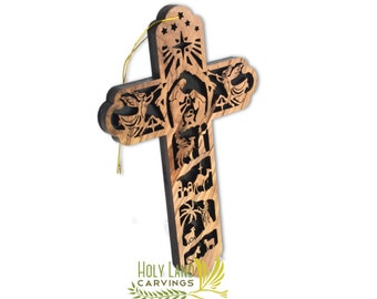 Puede incluir: Un adorno de cruz de madera con una talla detallada de la escena de la natividad. La cruz está hecha de madera oscura con un fondo negro y detalles dorados. El adorno tiene una cuerda dorada para colgar.