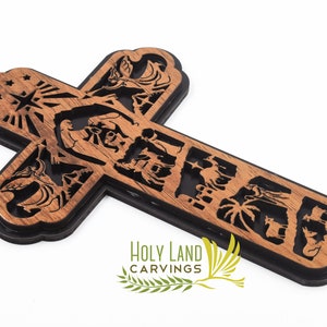 May include: A wooden cross with a black background, featuring intricate carvings of scenes from the Bible. The cross is decorated with a star and rays of light at the top, and the text "HOLY LAND CARVINGS" is visible at the bottom.