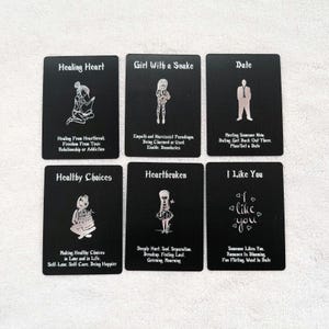 Può includere: Sei carte rettangolari nere con illustrazioni a linee bianche e testo. Le carte sono intitolate Healing Heart, Girl With a Snake, Date, Healthy Choices, Heartbroken e I Like You, ognuna con frasi descrittive sugli stati emotivi.