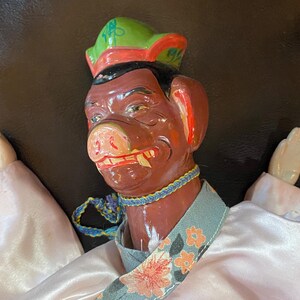 May include: A vintage Asian puppet with a pig-like face, wearing a green and red hat, a blue and yellow necklace, and a light blue floral collar. The puppet has white hands and a pink satin robe.