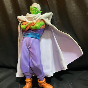 May include: A figurine of a green-skinned character with a white turban and cape. The character wears a purple outfit with a blue sash and orange boots. The figure has crossed arms and a determined expression.