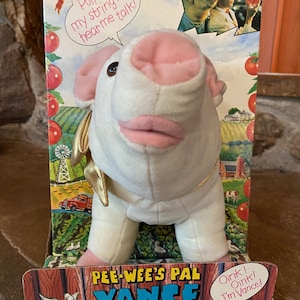 May include: A white plush pig with pink ears and a gold collar. The pig is wearing a white shirt and has a pull string on its back. The packaging says "Pee-wee's Pal Vance The Talking Pig" and "Oink! Oink! I'm Vance!"