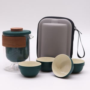 May include: A glass tea infuser set featuring a clear glass vessel with a dark green interior filter, a wooden lid, and a wooden band. The set includes three small, round tea cups with dark green exteriors and cream interiors, plus a grey carrying case.
