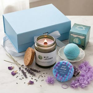 Może przedstawiać: Jasnoniebieskie pudełko prezentowe z białą wstążką, zapalona świeca Wild Lavender, niebieska kula do kąpieli, gąbka do mycia i gałązki lawendy. Świeca jest w szklanym słoiku z korkową pokrywką.