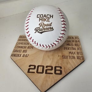 May include: A white baseball with red stitching featuring the text COACH MIKE Road Runners, resting on a light wood home plate plaque engraved with a list of names, jersey numbers, and the year 2026, including names like Cameron, Connor, and David.