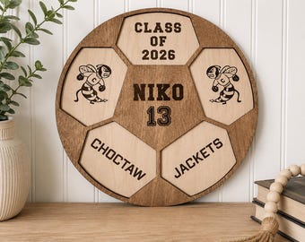 May include: A circular wooden sign shaped like a soccer ball with dark brown edges and light wood panels. It features the text "CLASS OF 2026", "NIKO", "13", "CHOCTAW", and "JACKETS" in dark brown, along with two illustrations of a mascot bee.