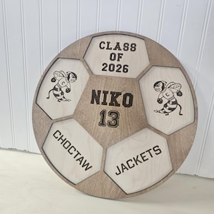 May include: A circular wooden sign shaped like a soccer ball with laser-engraved details. It features the text CLASS OF 2026, NIKO 13, CHOCTAW, and JACKETS, along with two illustrations of a cartoon bee mascot wearing boxing gloves.