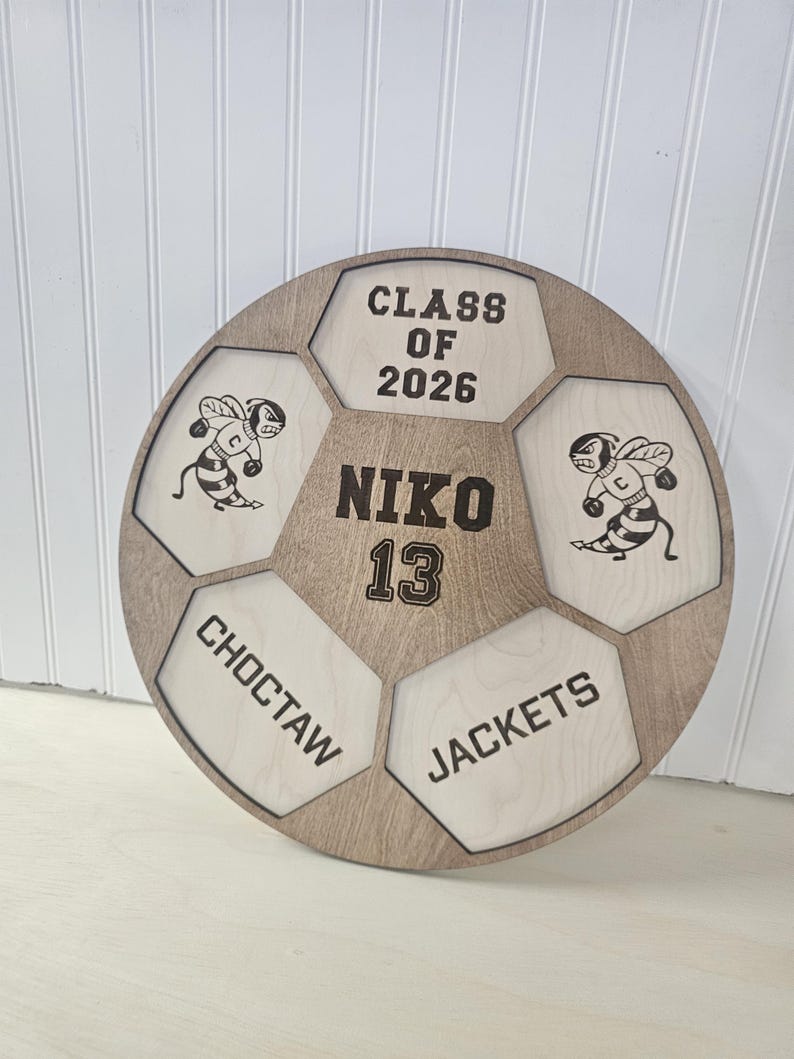 May include: A circular wooden sign shaped like a soccer ball with laser-engraved details. It features the text CLASS OF 2026, NIKO 13, CHOCTAW, and JACKETS, along with two illustrations of a cartoon bee mascot wearing boxing gloves.