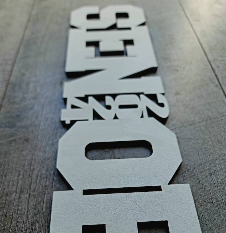 May include: White wooden letters spelling out the word "SENIOR" in a vertical stack. The letters are cut out and have a black outline.