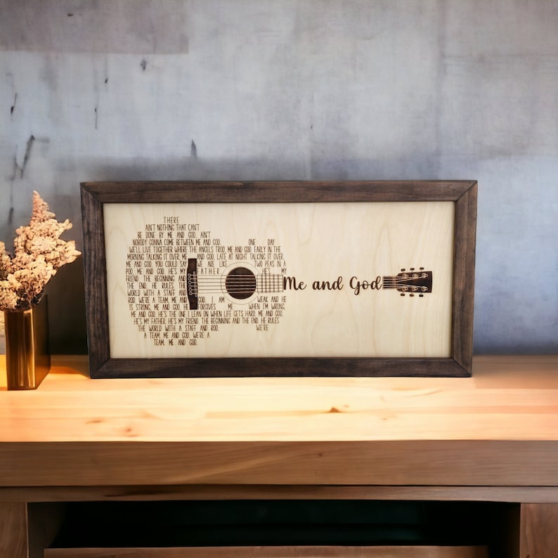 May include: A framed wood sign with a laser-cut design of a guitar and the lyrics to the song "Me and God". The lyrics are: "There ain't nothing that can't be done by me and God. Nobody gonna come between me and God. We'll live together where the angels trod. Me and God early in the morning talking it over. Me and God late at night talking it over. Me and God you could say we are two peas in a pod. Me and God He's my Father. The end He writes the world with a staff and rod. We were a team He and God. Is strong. We need each other. He forgives me when I'm wrong. He and God He's the one I lean on when life gets hard. Me and God. He's my father. He's my friend. The beginning and the end. He rules the world with a staff and rod. A team me and God. We were a team me and God."