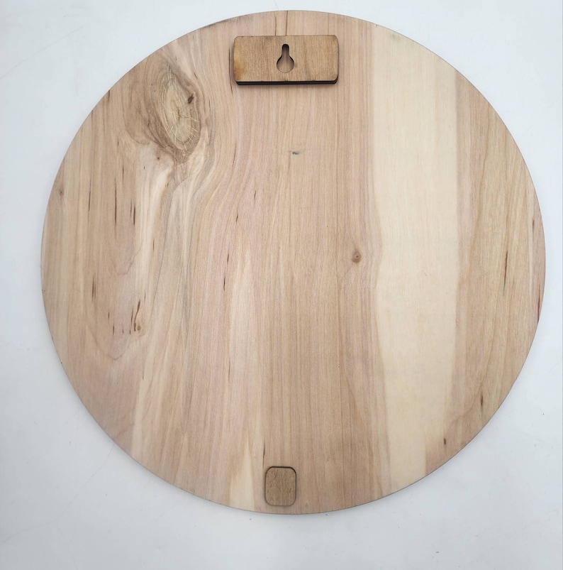 May include: A circular light-colored wooden plaque featuring a natural wood grain pattern with a visible knot. The back side includes a rectangular mounting bracket with a keyhole slot at the top and a small square spacer at the bottom edge.