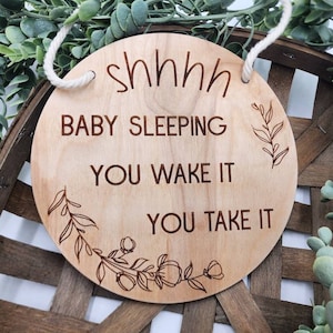 May include: A round wooden sign with the words "shhhh BABY SLEEPING YOU WAKE IT YOU TAKE IT" engraved in a dark brown font. The sign has a white rope for hanging and floral designs. The sign is displayed on a woven basket.