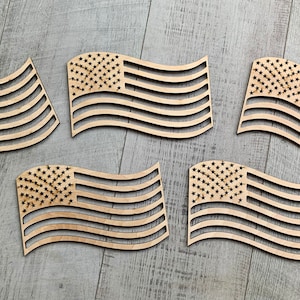 May include: Five wooden American flag cutouts, each with a wavy design, are arranged on a light wood surface. The flags are all the same size and shape, and they are all facing the same direction.