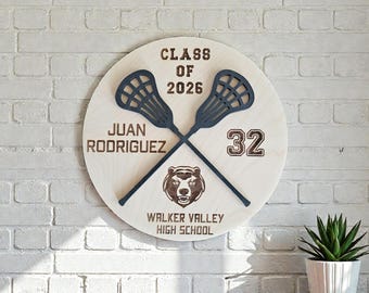 May include: A round wooden sign with the text "CLASS OF 2026" and "JUAN RODRIGUEZ 32" in brown lettering. Two crossed lacrosse sticks and a bear logo are also featured. The sign reads "WALKER VALLEY HIGH SCHOOL".
