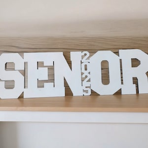 K&ouml;nnte beinhalten: Ein wei&szlig;es Holzschild mit der Aufschrift "SENIOR 2025" in Blockbuchstaben. Das Schild steht auf einem hellen Holztisch vor einem neutralen Hintergrund. Die Buchstaben sind verbunden und bilden ein zusammenh&auml;ngendes Design.