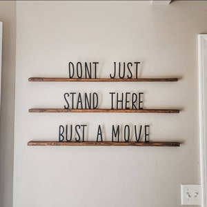May include: A wall-mounted decorative sign with three wooden shelves. The black text on the sign reads "DONT JUST STAND THERE BUST A MOVE". The shelves are mounted on a light-colored wall.