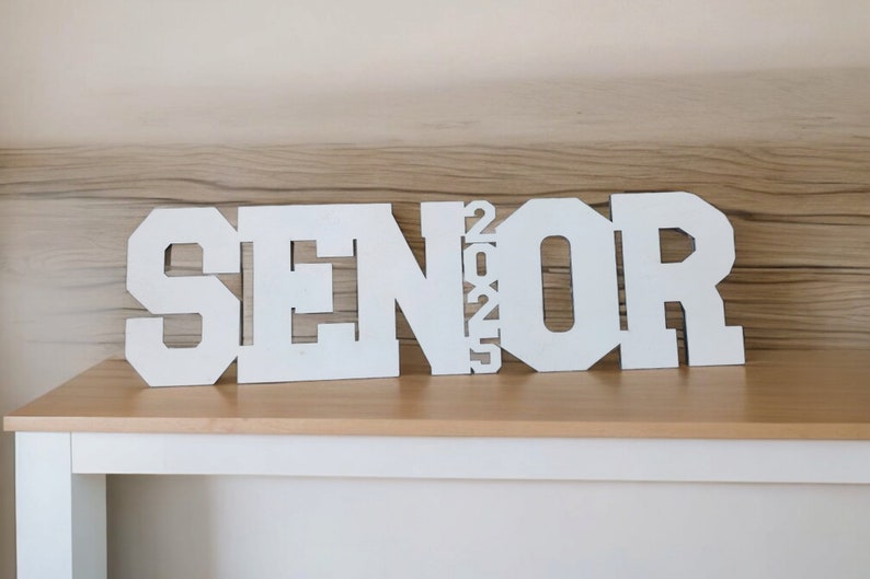 May include: White wooden letters spelling "SENIOR 2025" on a light brown wooden surface.