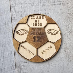 May include: A circular wooden plaque shaped like a soccer ball with laser-engraved details. It features the text CLASS OF 2025, SADIE JACKSON, 12, MARSH VALLEY, and EAGLES, along with two eagle head graphics in a light and dark wood finish.