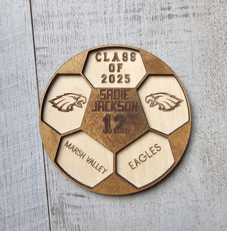 May include: A circular wooden plaque shaped like a soccer ball with laser-engraved details. It features the text CLASS OF 2025, SADIE JACKSON, 12, MARSH VALLEY, and EAGLES, along with two eagle head graphics in a light and dark wood finish.