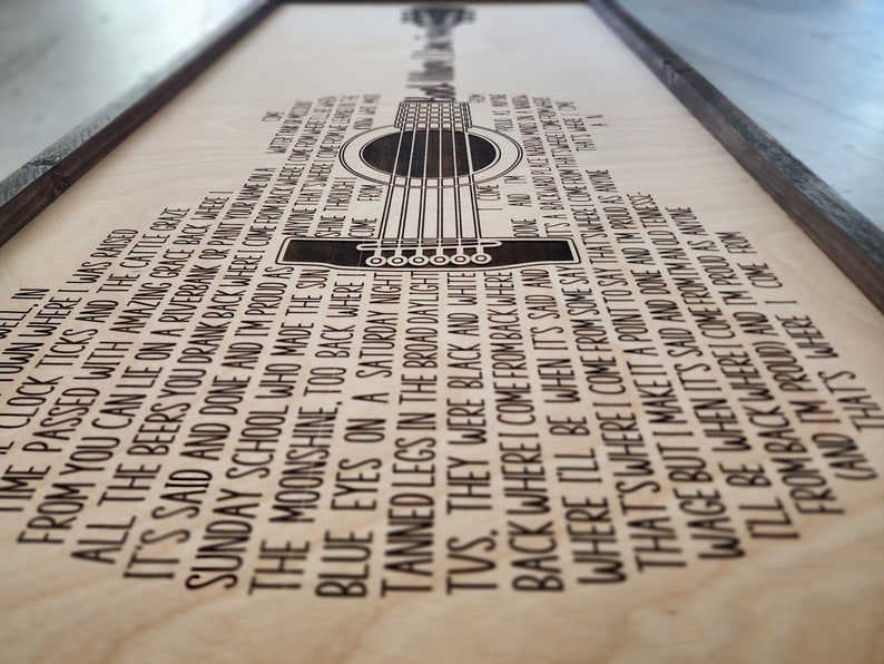 May include: A wood sign with a black and white silhouette of a guitar and lyrics from a song about a small town. The lyrics read: "Back where I was raised, clock ticks and the cattle craze, from you can lie on a riverbank or park, time passed with amazing grace back where it's said and done and I'm proud as all the beers you drank back where, Sunday school who made the sun and I'm proud as the moonshine, too back where, blue eyes on a Saturday night, tanned legs in the broad daylight, TVs, they were black and white, back where I come from and I'm proud as..."