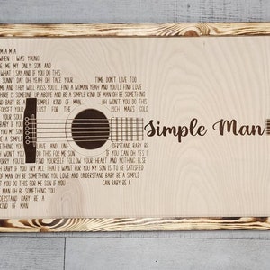 May include: A wooden sign with a burnt orange frame features the lyrics to the song "Simple Man" by Lynyrd Skynyrd. The lyrics are printed in a brown font and the sign includes a silhouette of an acoustic guitar.