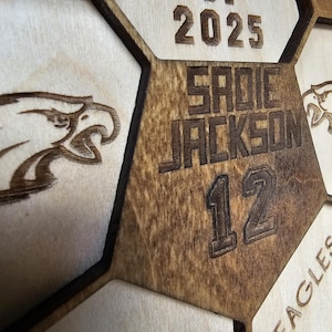 May include: A laser-cut wooden honeycomb display featuring the text CLASS OF 2025, the name SADIE JACKSON, and the number 12. The design includes engraved eagle head graphics and the word EAGLES, with dark brown stained edges and light wood surfaces.