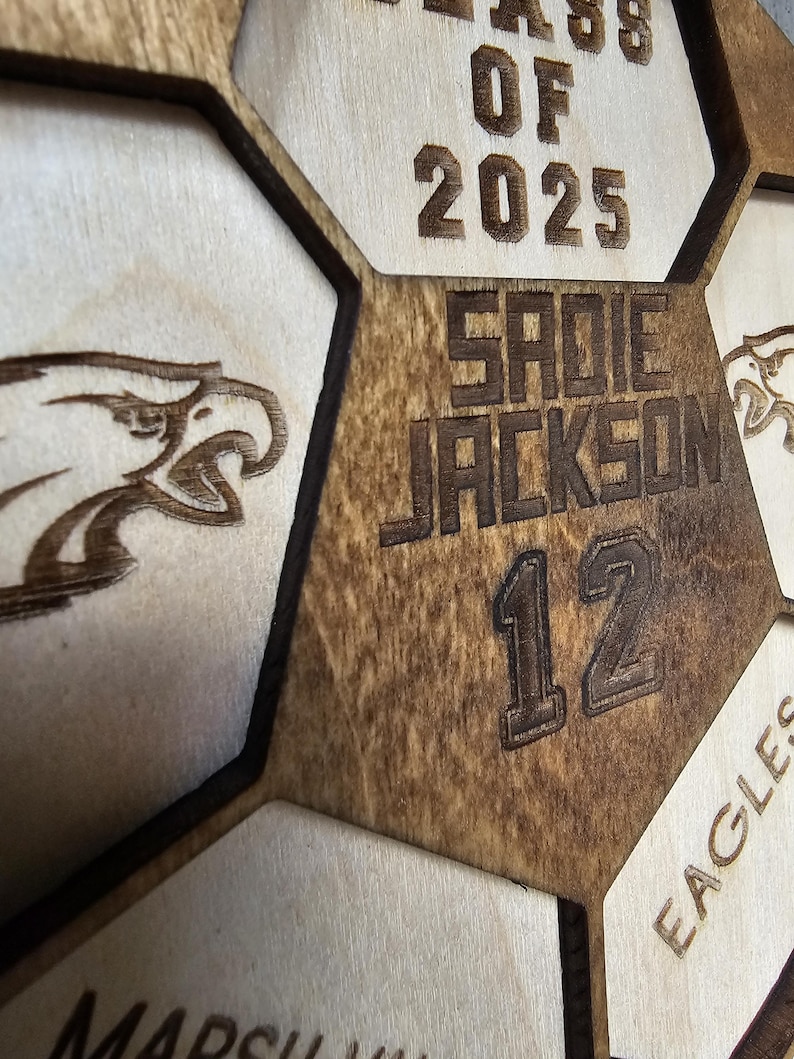 May include: A laser-cut wooden honeycomb display featuring the text CLASS OF 2025, the name SADIE JACKSON, and the number 12. The design includes engraved eagle head graphics and the word EAGLES, with dark brown stained edges and light wood surfaces.