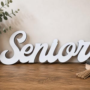 May include: A white cutout sign featuring the word Senior in a cursive script font. The letters have a smooth, matte finish with dark edges, creating a clean and bold appearance that stands out clearly against the surrounding neutral environment.