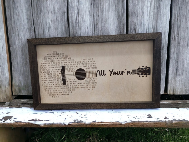 May include: A framed print with a brown wooden frame and a beige background. The print features a silhouette of an acoustic guitar with the lyrics to the song "All Your'n" by  the band,  "The Band Perry" printed on the background.