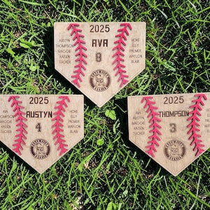 May include: Three wooden baseball home plate plaques with red stitching and personalized text. Each plaque features the year 2025, names, and a number. The Colorado Rockies logo is visible on each plaque. The plaques are on a bed of green grass.