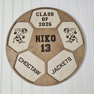 May include: A circular wooden wall sign shaped like a soccer ball with light and dark wood panels. It features the text CLASS OF 2026, NIKO, 13, CHOCTAW, and JACKETS, along with two engraved illustrations of a bee mascot wearing a letter C jersey.