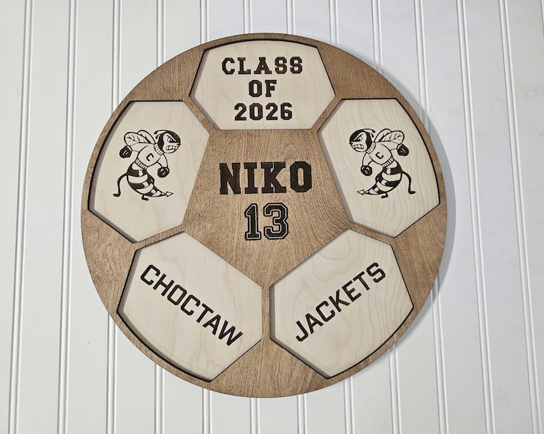 May include: A circular wooden wall sign shaped like a soccer ball with light and dark wood panels. It features the text CLASS OF 2026, NIKO, 13, CHOCTAW, and JACKETS, along with two engraved illustrations of a bee mascot wearing a letter C jersey.