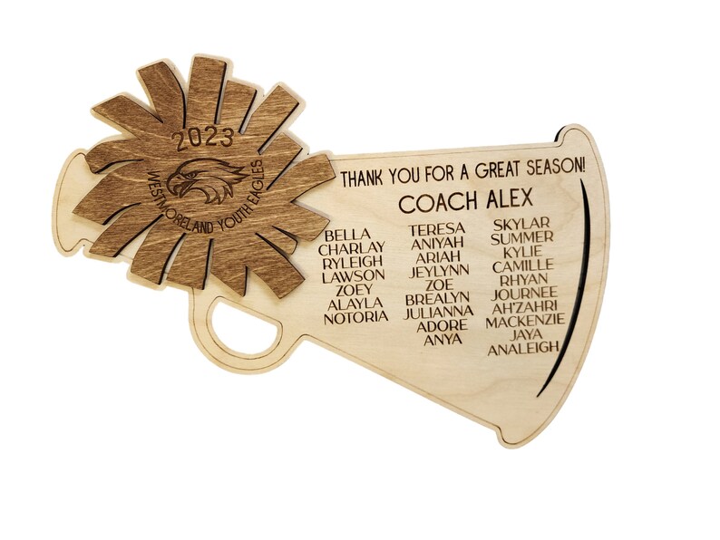 May include: A wooden megaphone with a brown wood grain finish. The megaphone has a starburst design on the top and the text "2023 Westmoreland Youth Eagles" on the front. The megaphone also has the text "Thank you for a great season! Coach Alex" and a list of names.